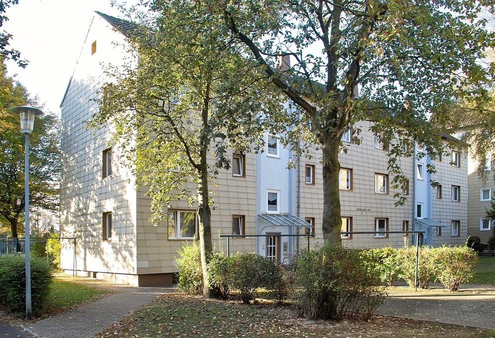 Mehrfamilienhaus mit Beige-Fassade, Bäumen und Gehwegen in Wohnanlage bei Tageslicht.