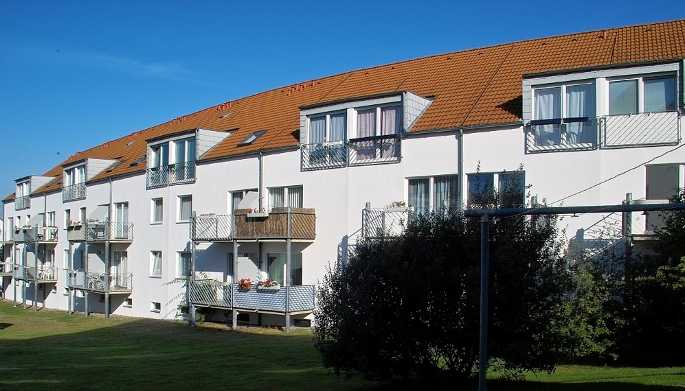 Mehrfamilienhaus mit weißen Wänden, roten Ziegeldächern und Balkonen vor blauem Himmel.