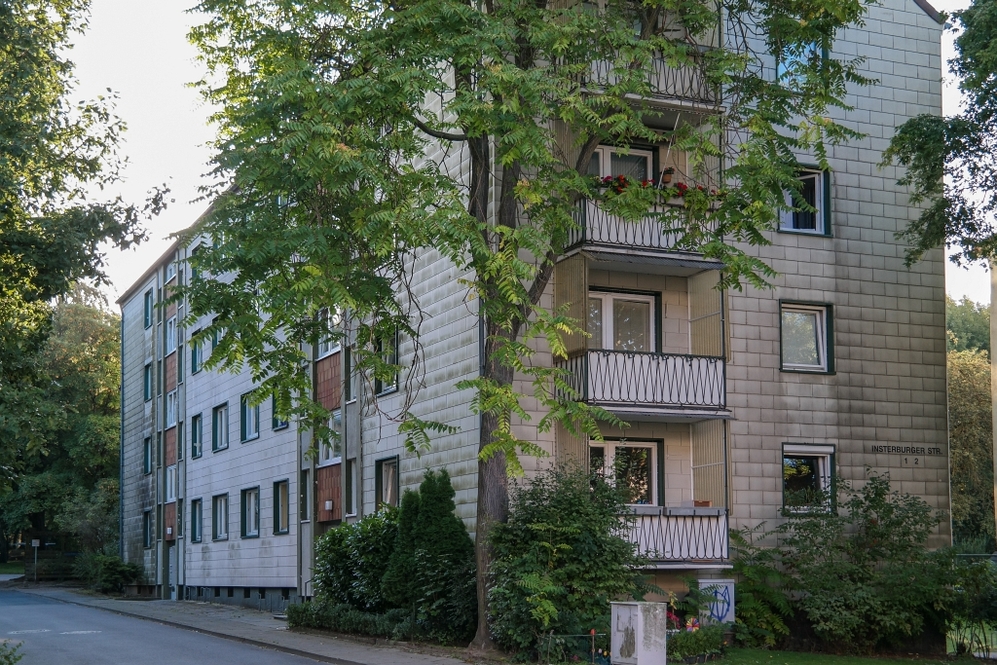 Mehrstöckiges Wohngebäude mit Balkonen, umgeben von Bäumen und Grünflächen an einer Straße.