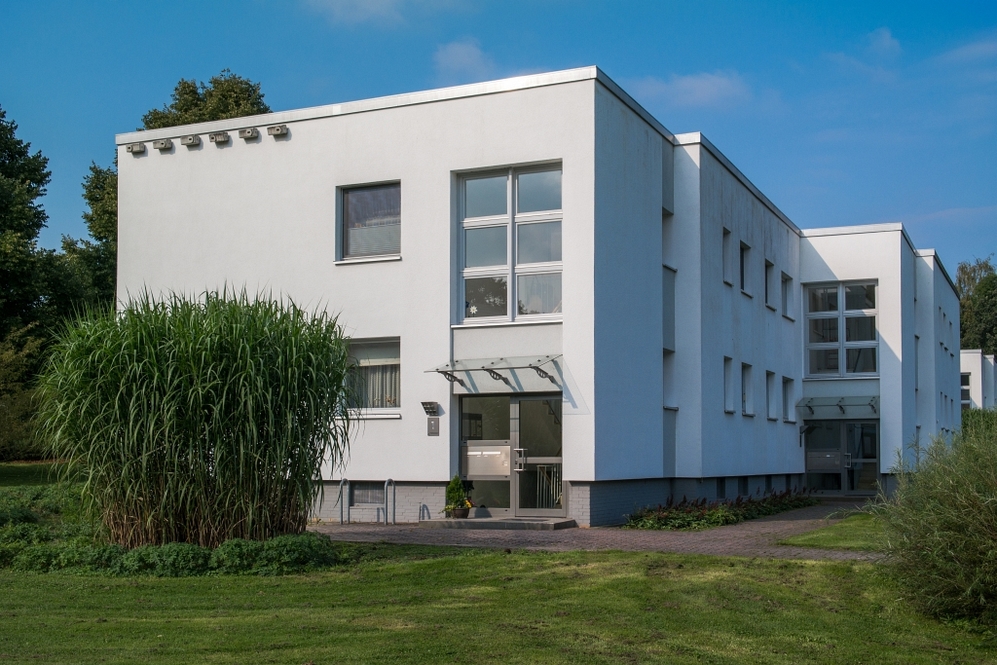 Moderner weißer Wohnkomplex mit großen Fenstern, Eingangsbereich und Grünfläche.