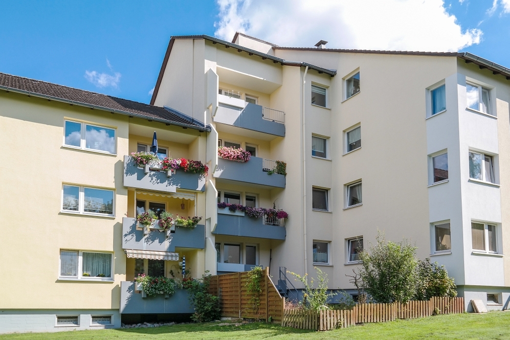 Mehrstöckiges Wohngebäude mit Balkonen und Blumen, umgeben von Grünfläche und Zaun.