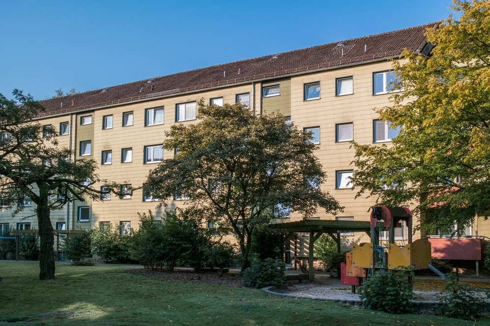 Mehrstöckiges Wohngebäude mit Spielplatz und Bäumen im grünen Innenhof bei Tageslicht.