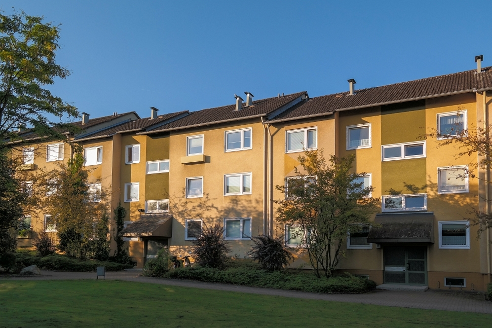 Mehrstöckiges Wohngebäude mit gelb-grüner Fassade und begrüntem Vorgarten bei Sonnenschein.
