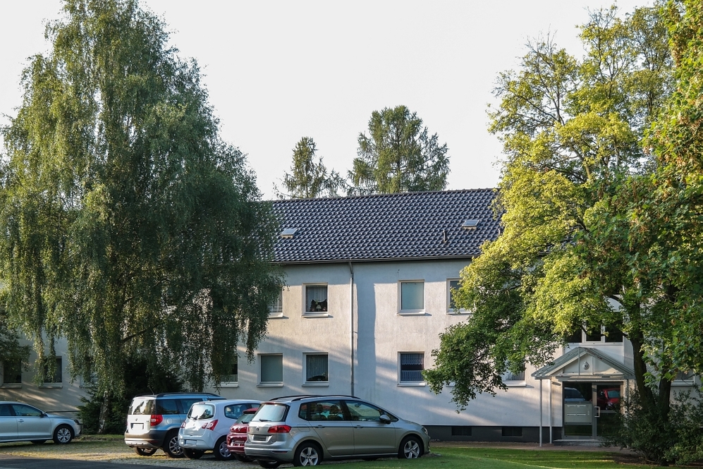 Mehrfamilienhaus mit Parkplätzen, umgeben von großen Bäumen und Grünfläche.