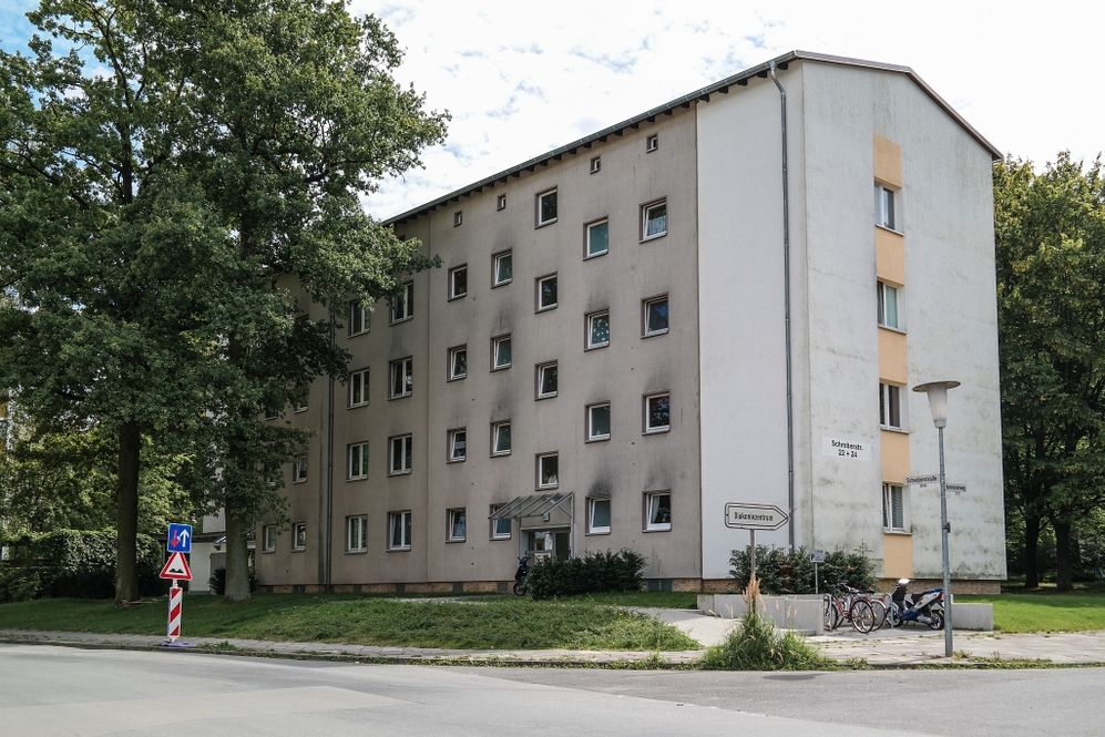 Mehrstöckiges Wohngebäude mit mehreren Fenstern und grünem Umfeld an einer Straßenecke.